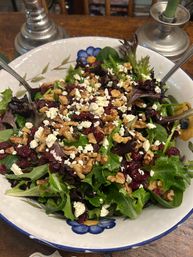Close-up of mixed green salad with baby spinach and red leaf lettuce, topped with crumbled feta, dried cranberries and chopped walnuts in a floral ceramic bowl on a wooden table