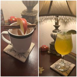 Cozy nightstand scene with a white mug of apple cinnamon tea garnished with cinnamon sticks and an apple slice, beside a sparkling citrus cocktail with lime in a wine glass under a vintage beaded lamp.