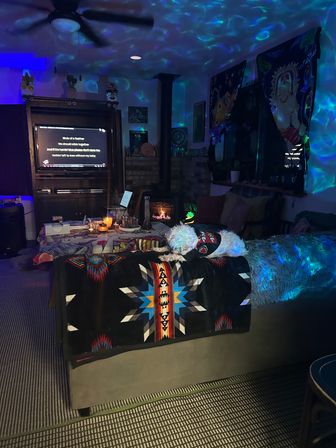 Cozy living room at night bathed in blue-green projector lights, candlelit coffee table and wood stove, TV displaying lyrics, and a small white dog lounging on a colorful southwestern blanket.
