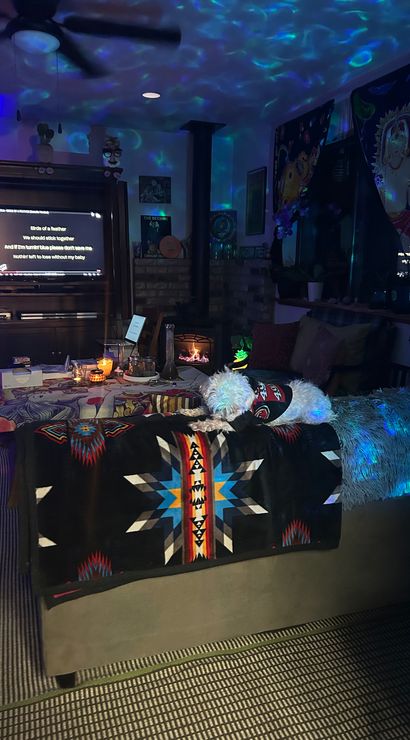 Cozy living room at night bathed in blue-green projector lights, candlelit coffee table and wood stove, TV displaying lyrics, and a small white dog lounging on a colorful southwestern blanket.