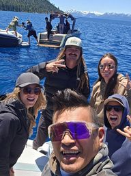 Smiling group taking a boat selfie on a deep-blue alpine lake with snow-capped mountains and nearby boats, friends in sunglasses and hats making playful hand signs.