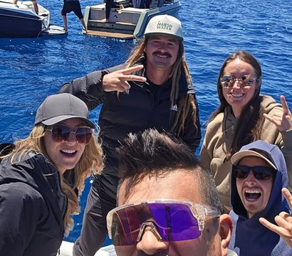 Smiling group taking a boat selfie on a deep-blue alpine lake with snow-capped mountains and nearby boats, friends in sunglasses and hats making playful hand signs.