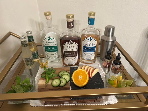 Home cocktail cart styled for mocktails: three clear non-alcoholic spirit bottles, metal shaker, ginger-ale bottles, sliced orange, apple, cucumber, lime wedges and fresh mint on a cutting board.