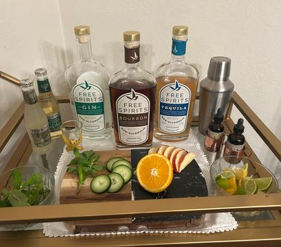 Home cocktail cart styled for mocktails: three clear non-alcoholic spirit bottles, metal shaker, ginger-ale bottles, sliced orange, apple, cucumber, lime wedges and fresh mint on a cutting board.