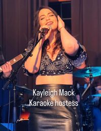 Female vocalist smiling into a microphone on a live-music bar stage during karaoke night, guitarist and drummer visible in the background.