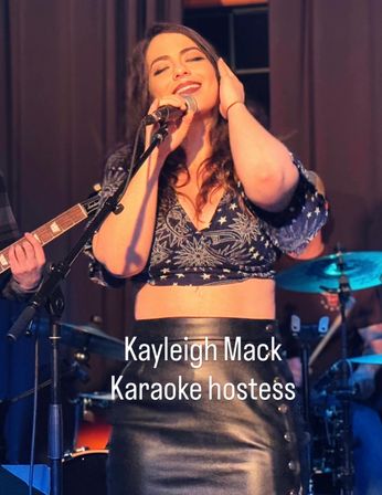 Female vocalist smiling into a microphone on a live-music bar stage during karaoke night, guitarist and drummer visible in the background.