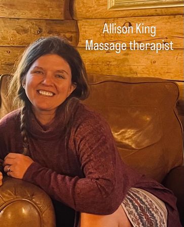 Smiling woman relaxing on a leather sofa in a cozy wood‑paneled cabin, wearing a maroon sweater and patterned skirt, with a visible 'Massage therapist' text overlay.