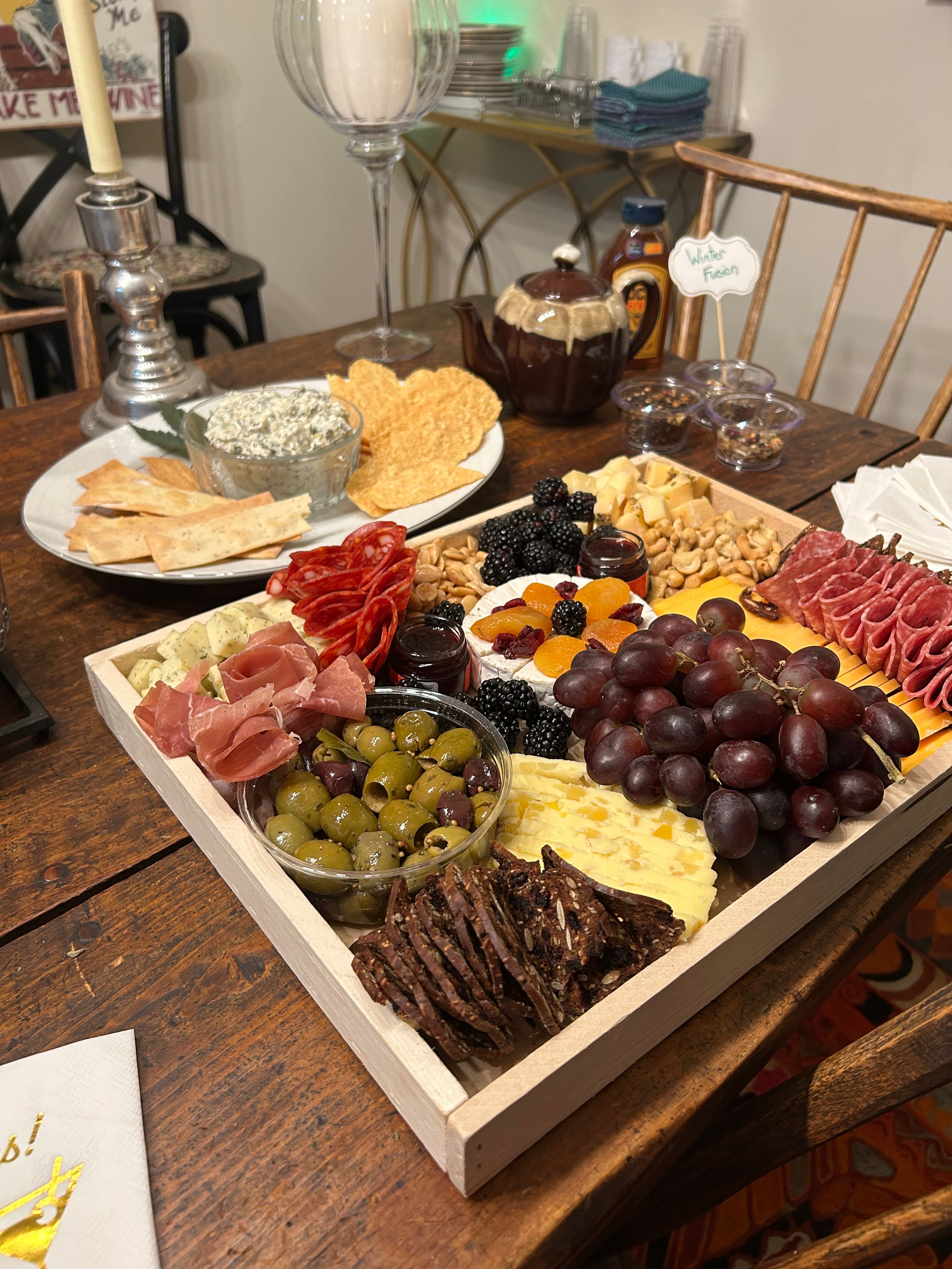 Close-up of a rustic charcuterie board on a wooden table with red grapes, blackberries, dried apricots, assorted cheeses, cured meats, mixed olives, nuts, crackers and jams, with a dip plate, candle and teapot in the background — a cozy appetizer spread.