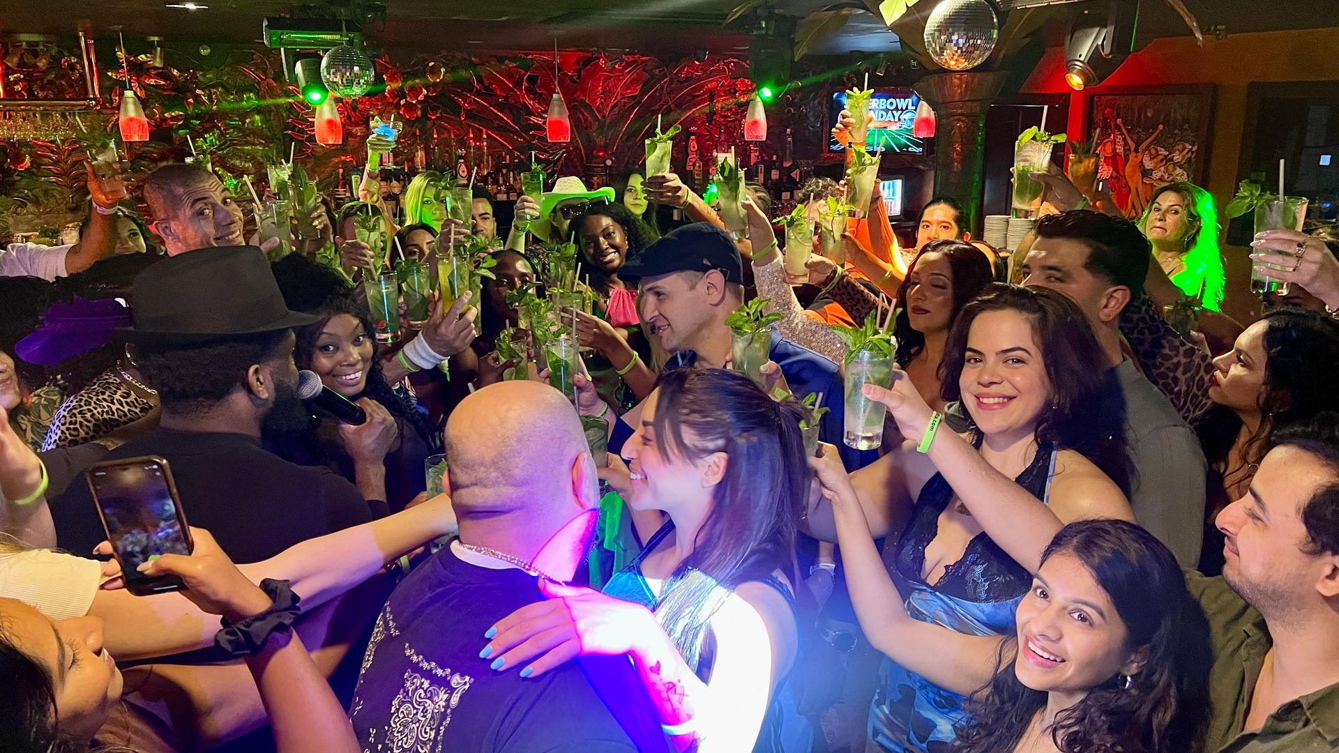Packed nightclub crowd cheering and raising mojito cocktails under colorful lights and a disco ball, singer with microphone and smiling friends celebrating together.