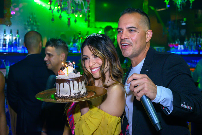 Smiling woman wearing a tiara holds a small chocolate drip birthday cake with lit candles on a platter while a man with a microphone celebrates beside her in a colorful nightclub bar.