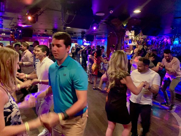 Crowded nightclub dance floor with couples dancing under purple and blue lights, lively bar and colorful decor in the background.