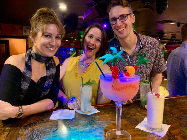 Three friends smiling at a lively cocktail bar, posing behind a wooden bar top with colorful tropical drinks — a large neon-pink tiki goblet with palm decorations and two frozen cocktails — under purple club lighting, nightlife scene.