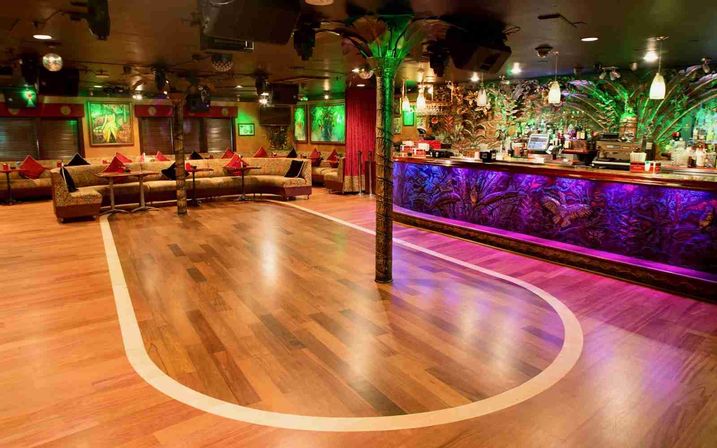 Tropical-themed nightclub interior with polished wooden dance floor outlined in white, curved lounge seating with red cushions, and a colorful illuminated bar featuring purple and green accent lighting.