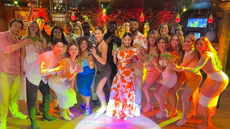 Large diverse group raising mojito cocktails at a tropical-themed bar, colorful party lighting and a woman with a microphone in the center — lively nightlife group photo.