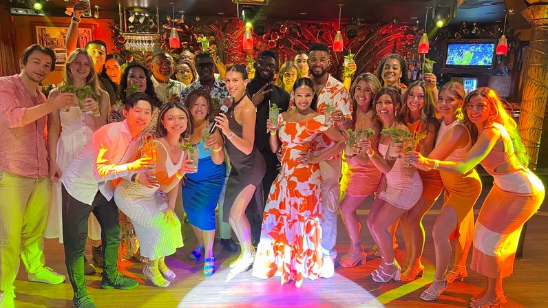 Large diverse group raising mojito cocktails at a tropical-themed bar, colorful party lighting and a woman with a microphone in the center — lively nightlife group photo.