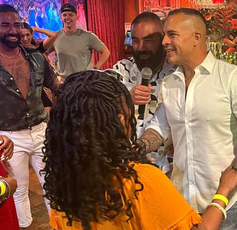 Nightlife scene at a lively bar — smiling group, man with a microphone and a man in a white shirt greeting a woman in an orange top under colorful red curtains.