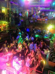 Vibrant two-level nightclub scene with a crowded dance floor, live brass player and singer on stage, neon purple-green lighting, balcony bar, hanging plants and patrons dancing