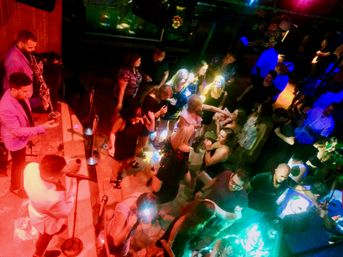 Aerial view of a crowded nightclub dance floor with a live singer and saxophonist on a small stage, people dancing and mingling under colorful red, green and blue party lights — lively urban nightlife scene.