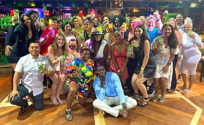 Group photo of diverse adults toasting with mint-garnished cocktails on a wooden dance floor inside a colorful bar, posing for a celebratory night out.