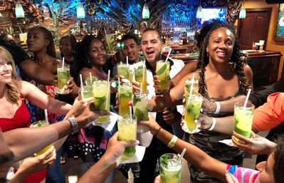 Group of people toasting with minty mojito-style cocktails at a lively indoor bar with colorful tropical decor and blue pendant lights