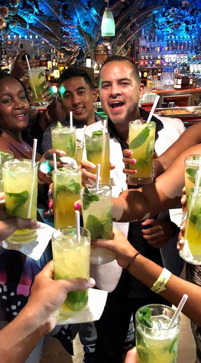 Group of people toasting with minty mojito-style cocktails at a lively indoor bar with colorful tropical decor and blue pendant lights