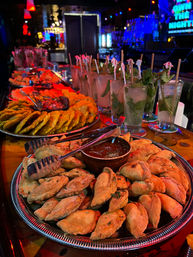 Neon-lit bar counter with silver platters of golden empanadas and dipping sauce beside a row of mint mojito cocktails, colorful late-night appetizer spread.