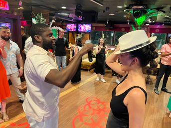 Man holding a microphone dances with a woman wearing a white fedora on a wooden dance floor while friends laugh and clap in a colorful indoor lounge with neon lights.