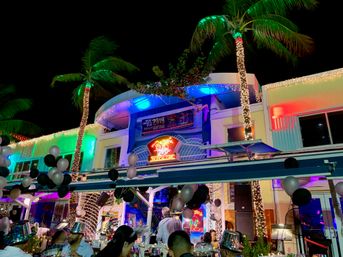 Festive tropical outdoor dining scene at night — palm trees wrapped in string lights, colorful neon signage, black-and-silver balloons and a New Year’s Eve crowd enjoying lively beachfront nightlife.
