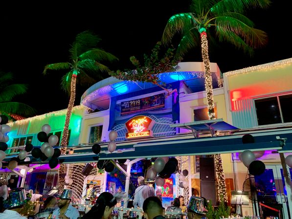 Festive tropical outdoor dining scene at night — palm trees wrapped in string lights, colorful neon signage, black-and-silver balloons and a New Year’s Eve crowd enjoying lively beachfront nightlife.