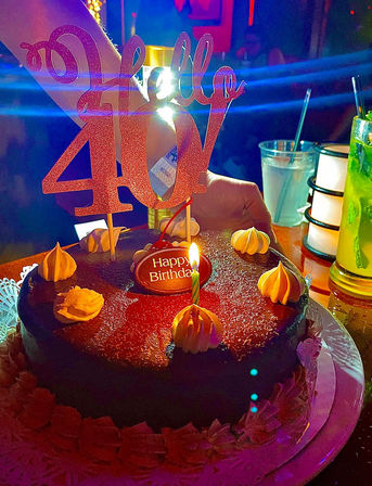 Chocolate 40th birthday cake with glittery "Hello 40" topper, single lit candle, piped cream dollops and a sparkling neon-lit bar table with cocktails nearby.