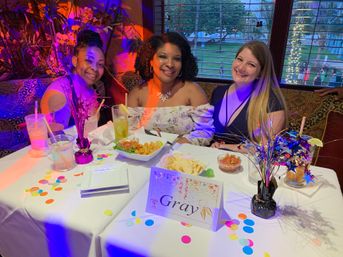 Three smiling women at a festive restaurant table with colorful confetti, starburst centerpieces, cocktails and appetizers on a white tablecloth, a tabletop card reading "Gray" and twinkling lights outside the window.