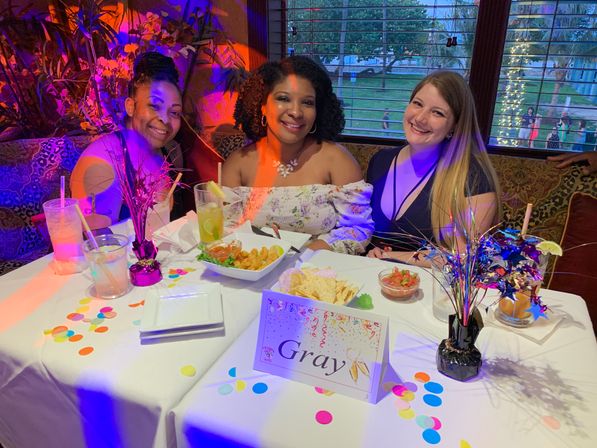 Three smiling women at a festive restaurant table with colorful confetti, starburst centerpieces, cocktails and appetizers on a white tablecloth, a tabletop card reading "Gray" and twinkling lights outside the window.