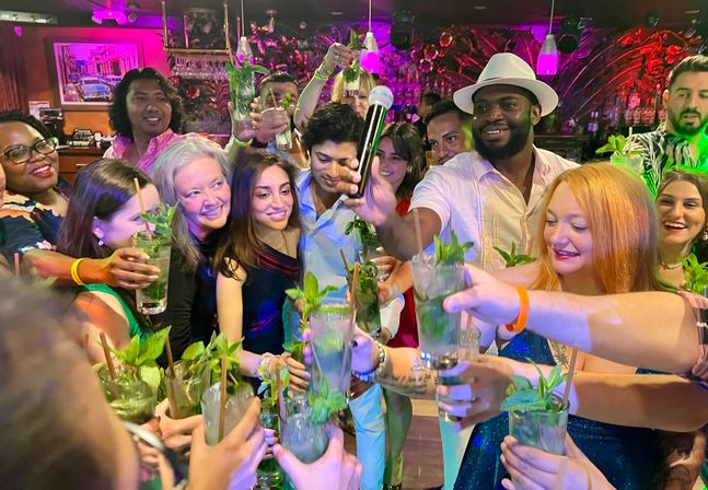Group of people toasting with mint mojito cocktails in a neon-lit bar, smiling around someone holding a microphone for a lively nightlife celebration