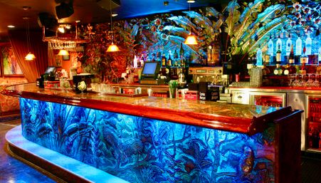 Vibrant tropical-themed cocktail bar interior with glowing blue carved bar front, polished wooden countertop, backlit liquor bottles on an ornate foliage mural, hanging pendant lights and bar equipment.