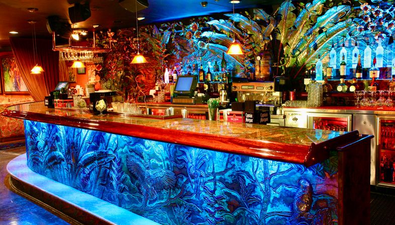 Vibrant tropical-themed cocktail bar interior with glowing blue carved bar front, polished wooden countertop, backlit liquor bottles on an ornate foliage mural, hanging pendant lights and bar equipment.