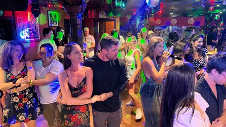 Lively crowded bar dance floor with couples dancing under colorful disco lights, a woman singing karaoke into a microphone, and patrons socializing in a festive nightlife scene.