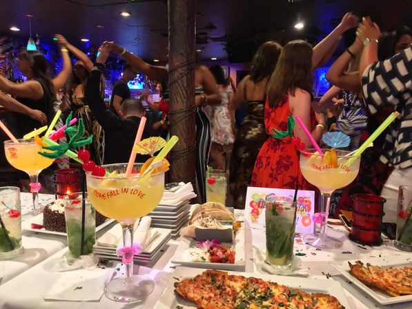 Lively dimly lit bar scene: colorful oversized tropical cocktails with umbrellas and neon straws on a table of pizza, tacos and desserts while a crowd dances in the background.