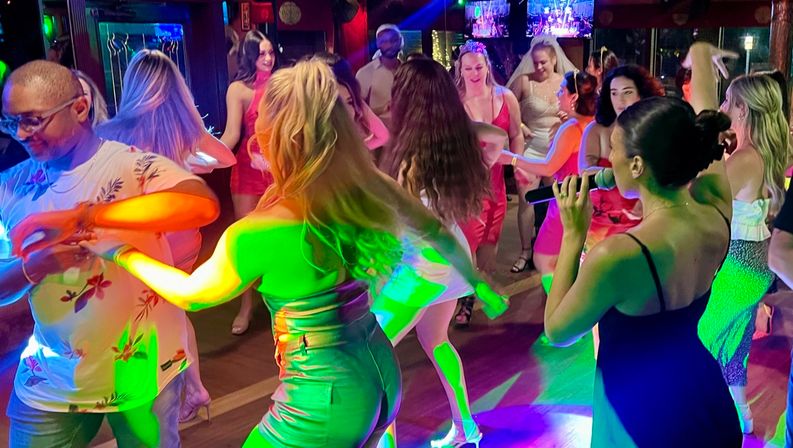 Lively nightclub dance floor with a singer on microphone and a mixed crowd in dresses and casual wear dancing under colorful party lights.