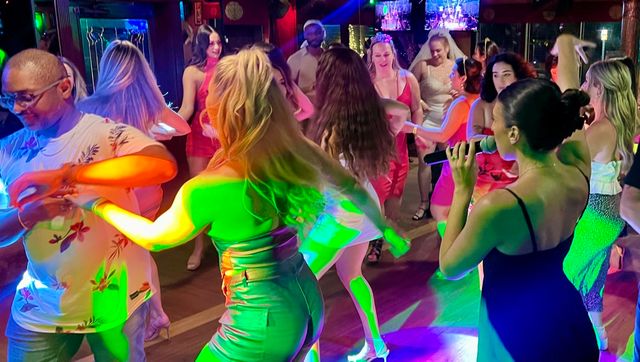 Lively nightclub dance floor with a singer on microphone and a mixed crowd in dresses and casual wear dancing under colorful party lights.