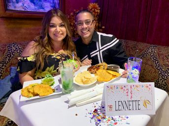 Two smiling diners in a cozy restaurant booth with plates of fried plantains, chips and grilled chicken, mojito-style cocktails, confetti and a festive place card on the table.