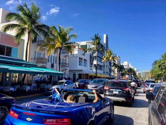 Sunny Miami Beach Ocean Drive scene with palm trees, white Art Deco hotels and sidewalk cafes, heavy traffic and a blue convertible with riders enjoying a day out.