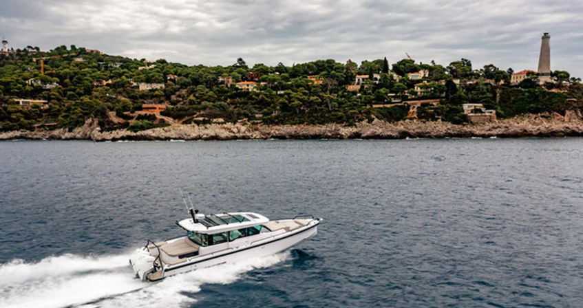 White cabin motorboat cutting through blue sea past a rocky, villa‑studded shoreline with trees and a tall lighthouse under a cloudy sky