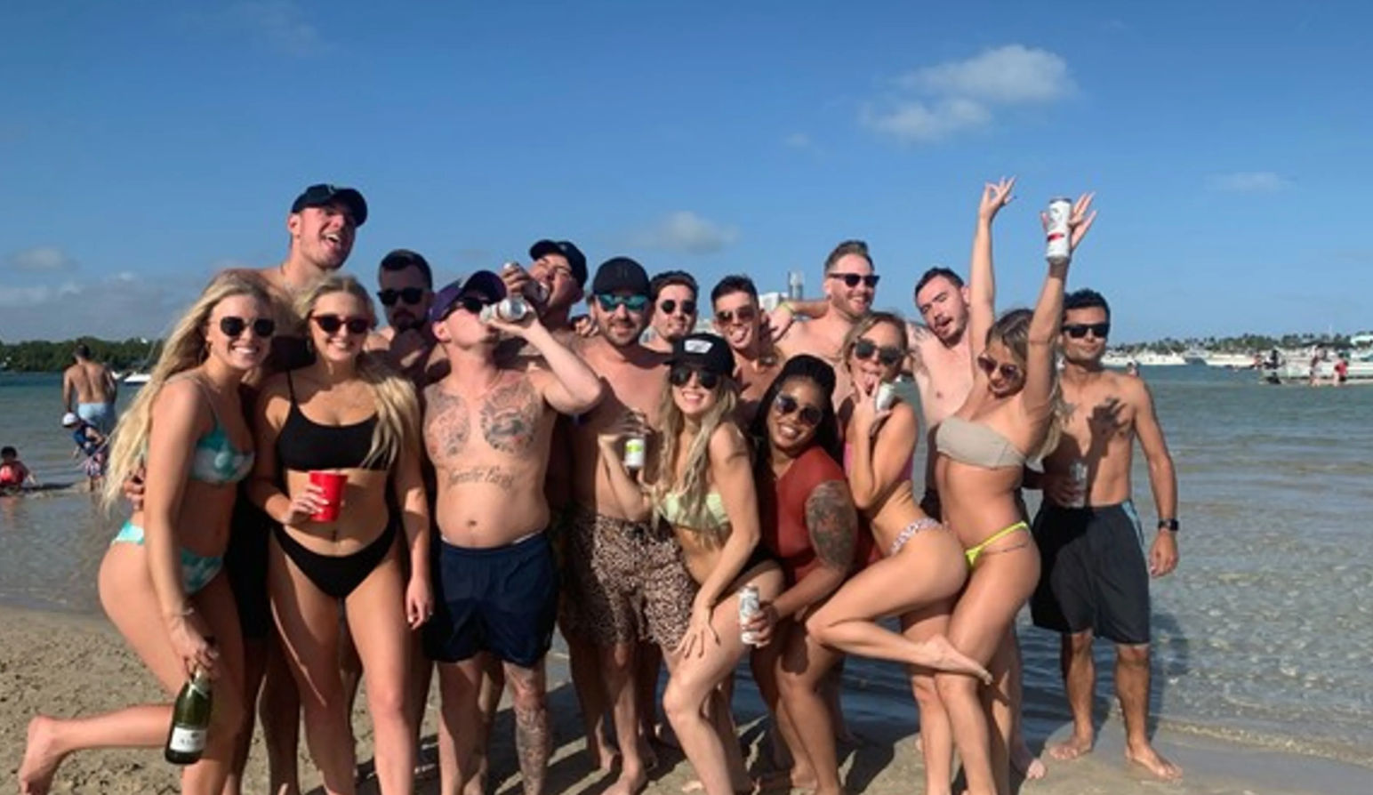 Group of young adults in swimwear posing and laughing on a sunny sandy beach by shallow blue water with anchored boats in the background, holding drinks and wearing sunglasses.