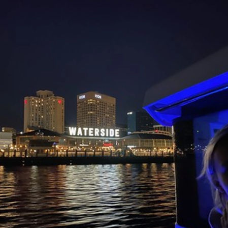 Nighttime urban waterfront scene with a large illuminated waterfront sign, hotel towers, a blue-lit boat edge in the foreground, and shimmering reflections on rippling water.