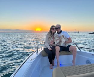 Sunset boat ride on the ocean with two people holding two small white dogs, sailboats dotting the horizon on a coastal cruise.