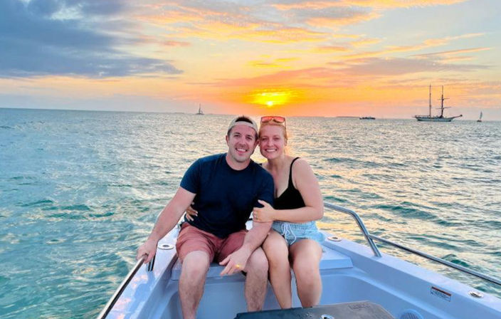 Smiling couple on a small boat at sunset over the ocean, sailboats and a tall ship on the horizon under a colorful sky