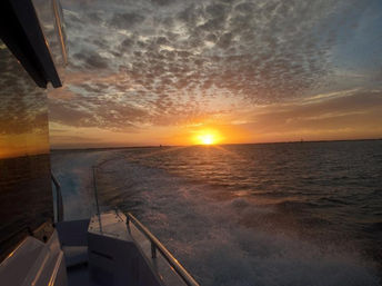 Golden sunset over open water seen from a boat's stern, frothy wake trailing toward the horizon, dramatic cloud-filled sky and sunset reflected on the boat's glossy side.