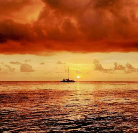 Fiery orange ocean sunset with a silhouetted sailboat on calm water beneath dramatic storm clouds — tropical seascape vista
