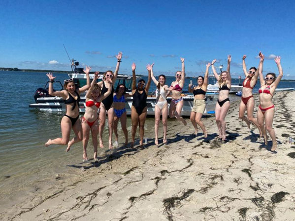 Group of friends jumping and cheering on a sandy beach beside a docked motorboat under a clear blue sky — lively summer bay shoreline scene in swimsuits.