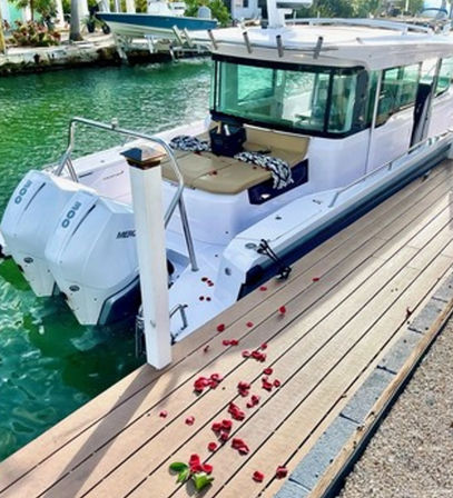 White cabin motorboat with twin outboards moored at a sunny tropical marina dock, turquoise water, tan cockpit cushions and scattered red rose petals on the wooden dock